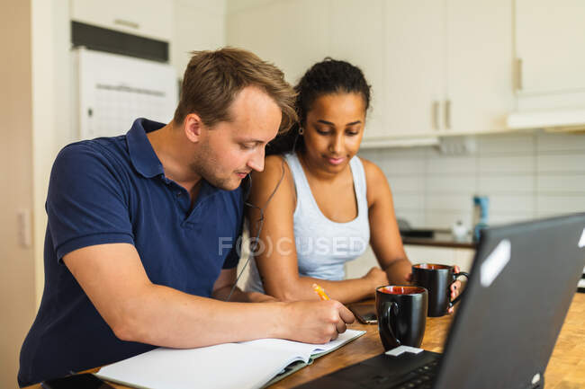 Sonriendo joven pareja multirracial utilizando aparatos y dispositivos mientras se sienta en el mostrador en la cocina y el estudio - foto de stock