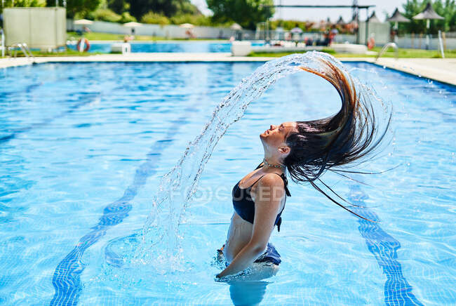 Side view of young female in swimwear standing in pool while splashing water and flicking hair — Stock Photo