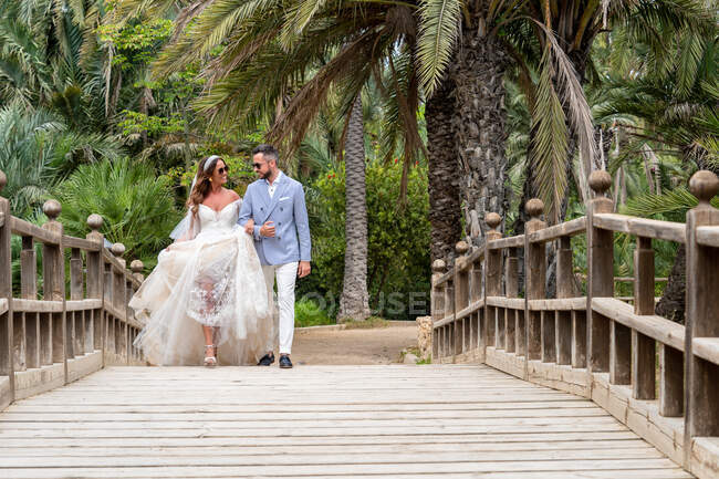 Супружеская пара в свадебных нарядах ходит по деревянному пешеходному мосту с перилами, держа за руки и глядя друг на друга возле зеленых пальм и растений в саду в летний день — стоковое фото