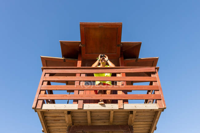 Baixo ângulo de olhar através de binóculos na torre de vigia de madeira enquanto supervisiona a segurança no mar contra o céu azul sem nuvens — Fotografia de Stock