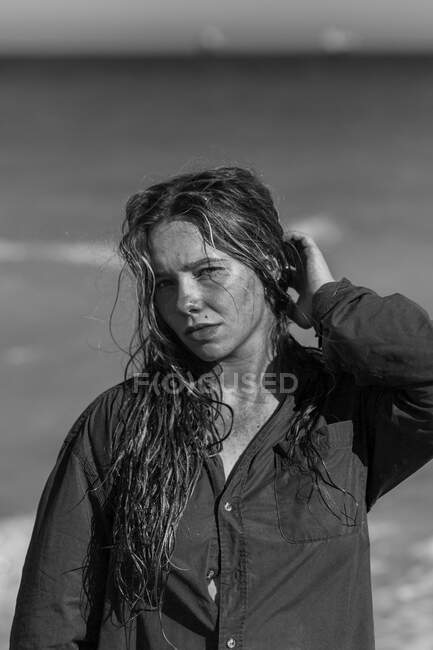 Bianco e nero di femmina in camicia bagnata e con i capelli bagnati in piedi guardando la fotocamera sulla spiaggia vicino al mare mentre si gode la giornata estiva — Foto stock
