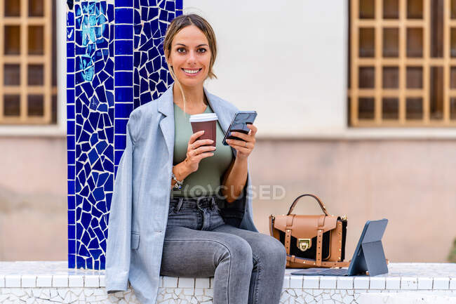 Donna positiva in abito elegante messaggistica di testo sul cellulare e guardando la fotocamera mentre seduto con caffè da asporto su panca con borsa e tablet — Foto stock
