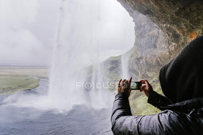 Side view of unrecognizable traveler in warm outerwear and hoodie taking photo of picturesque Seljalandsfoss waterfall on smartphone during trip in Iceland — Stock Photo