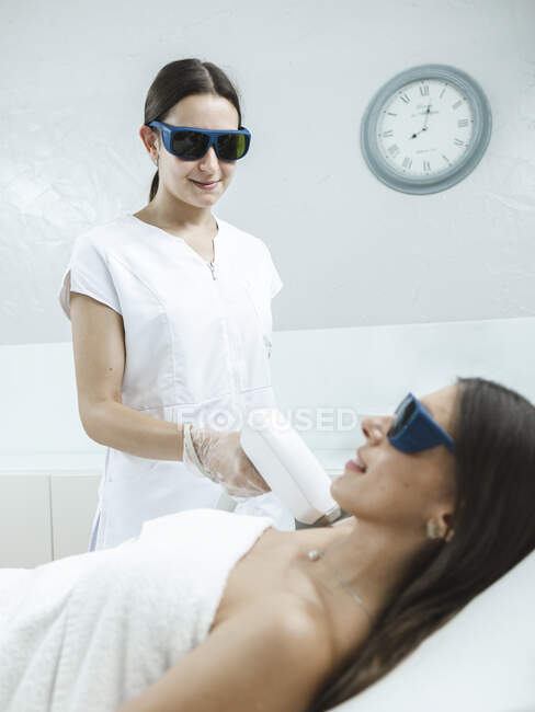 Jeune maître en lunettes faisant la procédure d'épilation au laser avec un équipement professionnel à la main dans le salon de lumière — Photo de stock