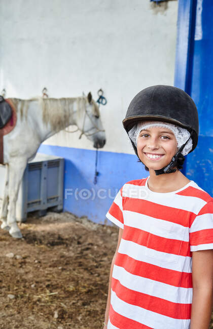 Усміхнена дитина в шапці для хокею і повсякденному одязі, дивлячись на камеру, стоячи біля білого коня в стайні біля стіни будівлі в денний час — стокове фото