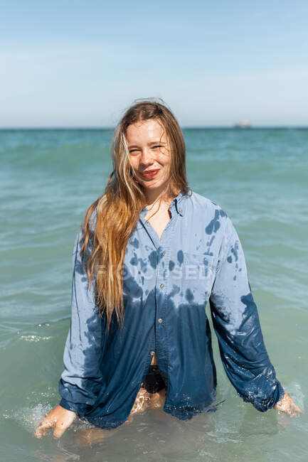 Encantadora joven hembra feliz en camisa mojada de pie en el agua de mar y mirando a la cámara en el día soleado de verano - foto de stock