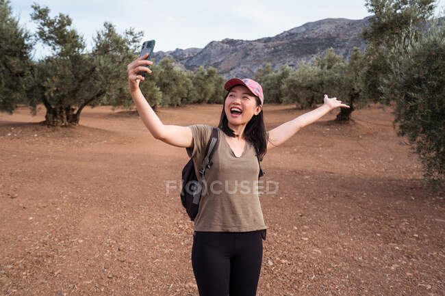 Delighted Asian female traveler with spread arms taking self portrait on smartphone while standing on plantation with olive trees in countryside — Stock Photo