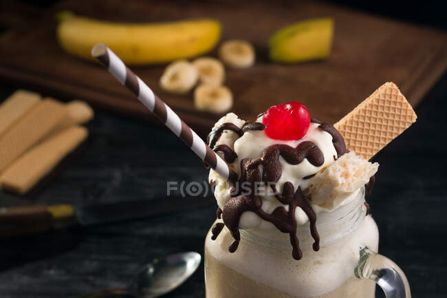 Du dessus du bocal en verre de milkshake à la banane sucrée garni de gaufres à la crème fouettée chocolat et cerise sur la table — Photo de stock