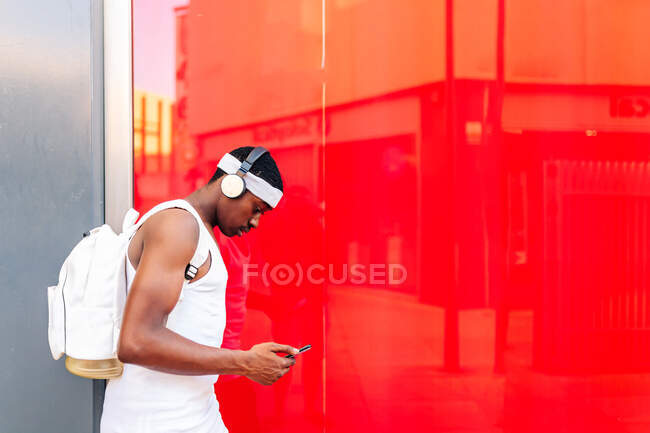 Side view African American male in white clothes listening to music in wireless headphones and messaging on mobile phone while walking against red wall in city — Stock Photo