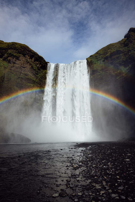 Захватывающий дух пейзаж радуги, сияющей над быстрым мощным водопадом Скогафосс, протекающим через скалистую скалу в Исландии — стоковое фото