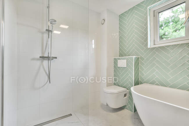 Cabine de chuveiro de vidro com mangueira colocada perto da banheira branca e WC em luz espaçosa casa de banho com janela de vidro no apartamento — Fotografia de Stock