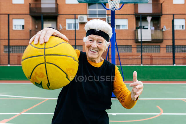 Mulher madura positiva em activewear e headband olhando para a câmera enquanto está de pé com bola no braço estendido durante o jogo de basquete — Fotografia de Stock
