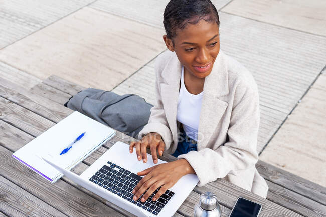 Зверху позитивна афро-американська жінка-фрилансер переглядає нетбук, сидячи на вулиці за записником під час онлайн-роботи. — стокове фото