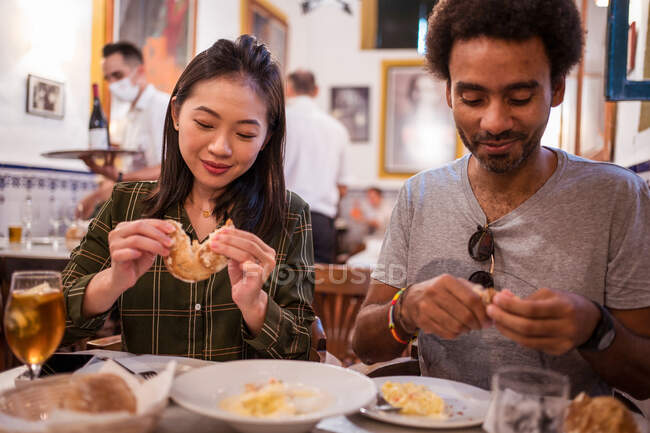 Feliz joven pareja multiétnica en ropa casual sonriendo mientras come deliciosos alimentos durante la fecha en el restaurante moderno - foto de stock