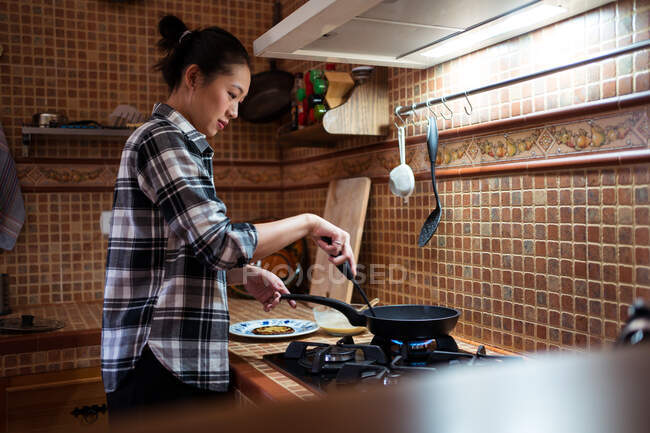 Бічний погляд на сфокусовану молоду азіатку в повсякденному одязі куховарські млинці з використанням спатули, стоячи на кухні. — стокове фото
