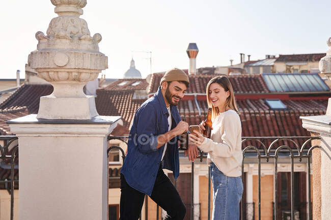 Side view of positive young friends using cellphone together while drinking beer from glass bottles and leaning on fence on balcony — Stock Photo