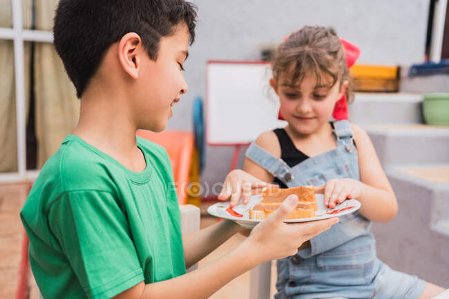 Niños contentos en ropa casual con plato de pan fresco con mermelada dulce en sala de luz con pizarra blanca en casa - foto de stock