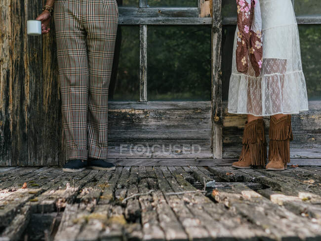 Two anonymous people, he in plaid pants and she in a see-through dress and boots standing on the porch of an abandoned wooden house — Stock Photo