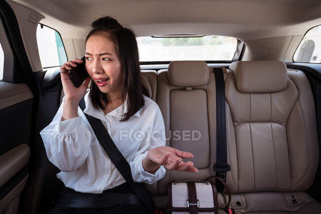 Passageiro étnico confuso com cinto de segurança apertado falando no celular enquanto andava no banco de trás em táxi — Fotografia de Stock