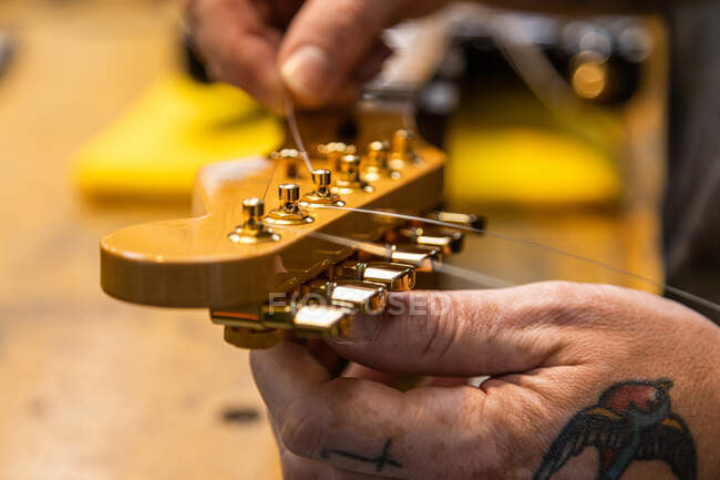 Crop unrecognizable musician with tattoos on hand changing strings on guitar in workshop — Stock Photo