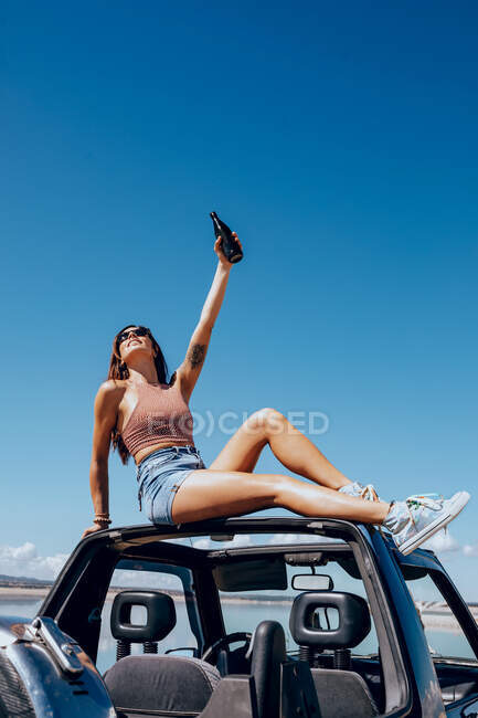 Full body of happy young female in summer outfit and sunglasses raising hand of beer while sitting on roof of safari car on shore of river — Stock Photo