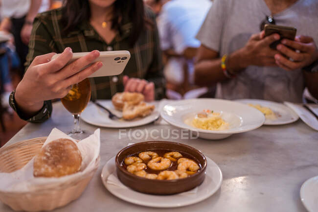 D'en haut de la culture femme anonyme prenant des photos de délicieux aliments avec des crevettes souriantes sur smartphone tout en dînant avec petit ami au restaurant — Photo de stock