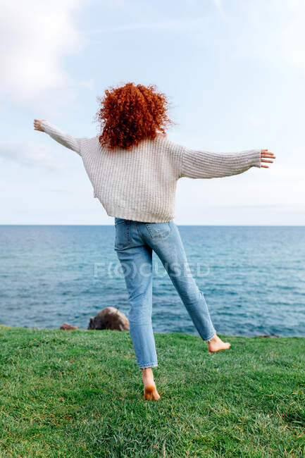 Vista trasera de cuerpo completo de irreconocible mujer de pelo rizado feliz extendiendo los brazos mientras disfruta de la libertad en la colina cubierta de hierba en la costa del mar - foto de stock