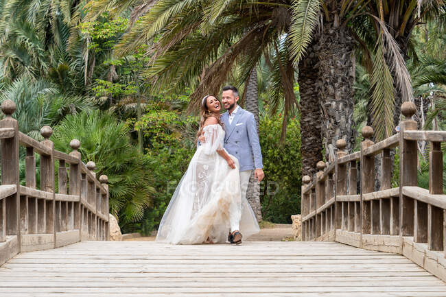 Супружеская пара в свадебных нарядах, стоящая на деревянном пешеходном мосту с перилами, обнявшись и глядя на зеленые пальмы и растения в саду в летний день — стоковое фото