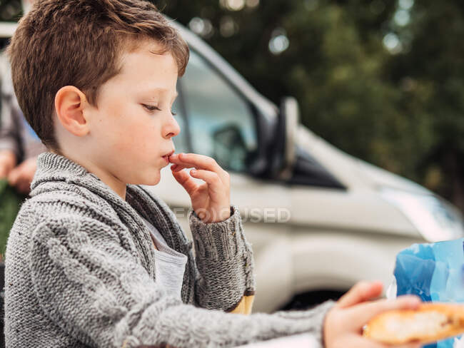 Side view of crop adorable little boy in warm knitted jacket eating snacks sitting near car parked in nature during road trip — Stock Photo