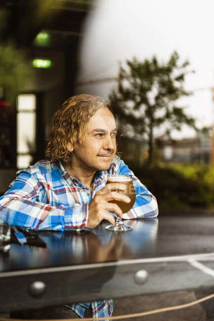 Macho pensativo com cabelo encaracolado em roupas casuais sentado no balcão de madeira perto da janela no bar e bebendo cerveja durante o dia — Fotografia de Stock