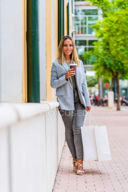 Vista frontale di una donna positiva in abito elegante con caffè per andare a guardare la fotocamera mentre tiene in mano un paio di borse della spesa — Foto stock