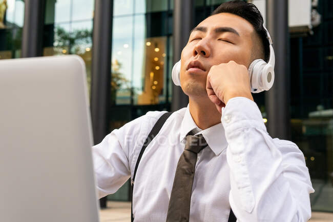 Mindful jeune entrepreneur asiatique avec les yeux fermés profiter de la chanson des écouteurs sans fil contre ordinateur portable dans la cafétéria de la rue — Photo de stock