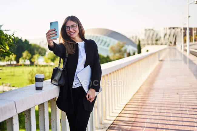 Donna positiva in elegante abito formale in piedi con computer portatile e prendendo autoritratto sul cellulare vicino ringhiera del ponte — Foto stock