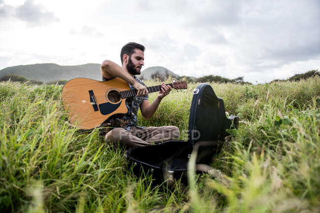 Calma musicista maschile seduto con chitarra acustica su erba verde in campo contro collina sotto cielo nuvoloso di giorno — Foto stock