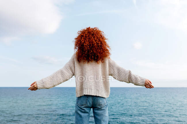 Back view of unrecognizable delighted female with curly long ginger hair raising hands on shore of rippling sea — Stock Photo