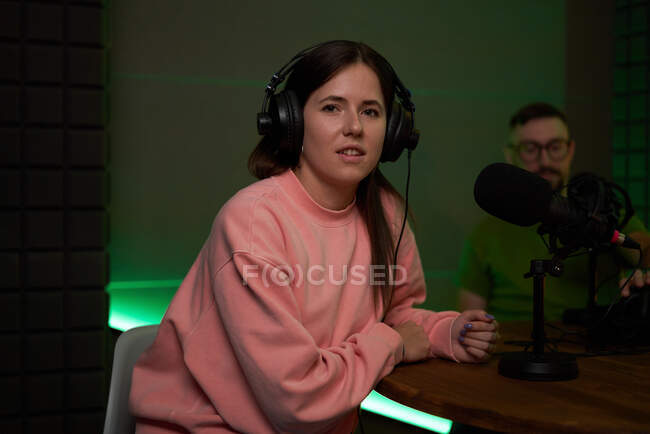 Positive young female radio host in casual clothes and headphones smiling and looking at camera while recording podcast with male coworker in dark studio — Stock Photo