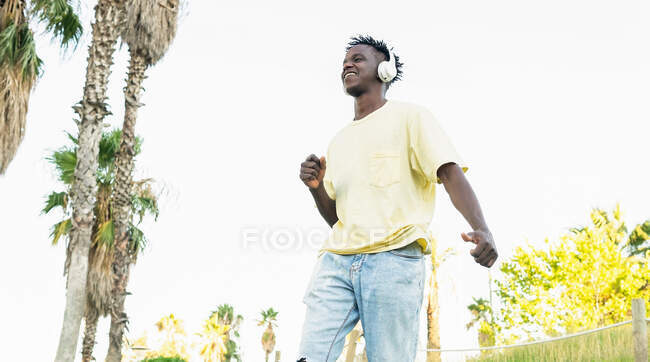 Знизу, у випадковому одязі афроамериканського юнака, який слухає музику навушниками і дивиться в парк у сонячний день. — стокове фото