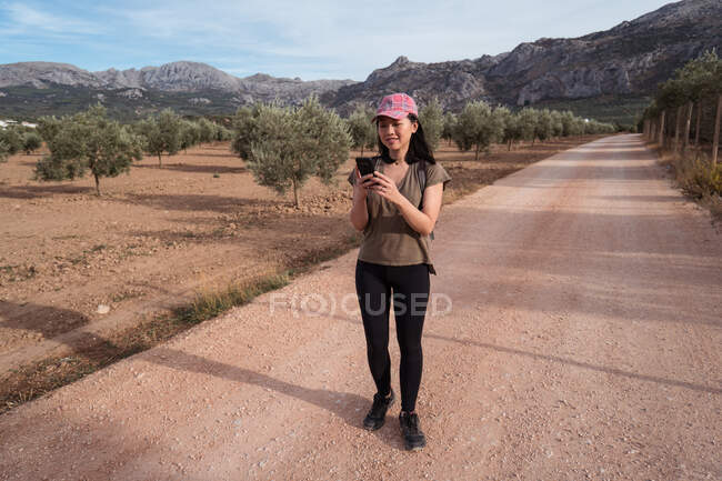 Full body of content Asian female traveler surfing cellphone while standing on footpath near plantation with lush green olive trees — Stock Photo