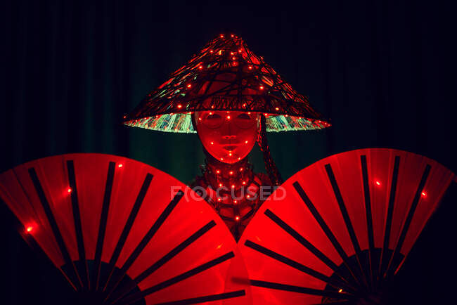 Mujer irreconocible con una máscara en traje tradicional creativo y ropa vietnamita con iluminación roja de pie en un estudio oscuro sobre fondo negro durante la actuación - foto de stock