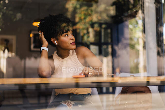 Attraverso il vetro di sognante giovane donna afroamericana in abito elegante e auricolari TWS toccare i capelli ricci e distogliere lo sguardo mentre seduto al bancone in caffè moderno e utilizzando smartphone — Foto stock
