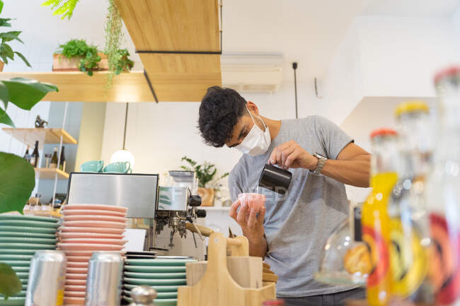 Ethnic barista in protective mask pouring milk into mug with coffee while standing at counter with coffeemaker and dishware in coffee house — Stock Photo