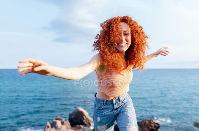 Feliz cabelo encaracolado fêmea estendendo os braços enquanto desfruta de liberdade na costa montanhosa da costa olhando para a câmera — Fotografia de Stock