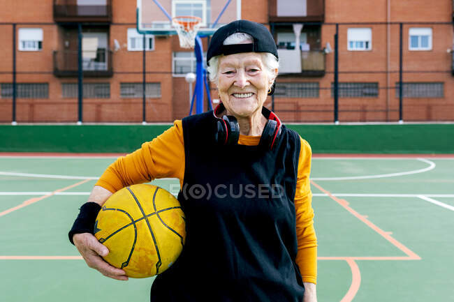 Ottimista donna matura in activewear e cuffie guardando la fotocamera mentre in piedi sul campo da basket pubblico con palla durante l'allenamento — Foto stock