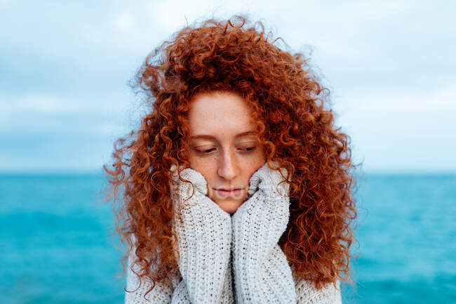 Femme immobile aux longs cheveux bouclés au gingembre portant un pull tricoté debout regardant vers le bas contre la mer bleue — Photo de stock