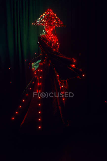Femme méconnaissable avec un masque en tenue traditionnelle créative et des coiffures vietnamiennes avec éclairage rouge debout en studio sombre sur fond noir pendant la performance — Photo de stock