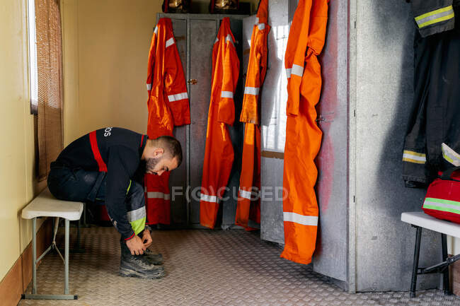 Cuerpo completo de hombre joven con uniforme de fuego y sentado en el banco en la estación de bomberos mientras ata cordones de zapatos cerca de un casillero personal durante el día. - foto de stock
