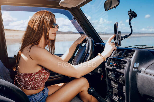 Vue latérale d'une jeune voyageuse confiante avec de longs cheveux roux en vêtements d'été en utilisant un navigateur sur téléphone portable tout en conduisant une voiture moderne près de la mer le jour ensoleillé — Photo de stock