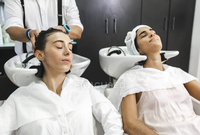 Молоді жінки в білому одязі закривають очі, миючи волосся в сучасному салоні краси — стокове фото