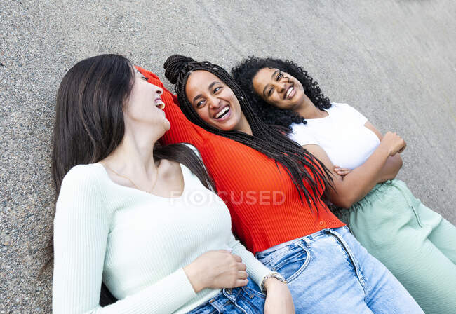 De cima de namoradas multirraciais alegres em roupas da moda rindo e deitado na estrada de asfalto na rua durante o dia — Fotografia de Stock