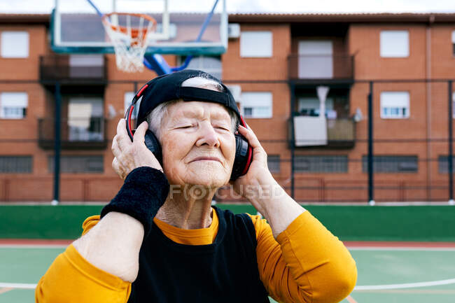 Content mature female with closed eyes in activewear listening to music with headphones while standing on public basketball court during training on street — Stock Photo
